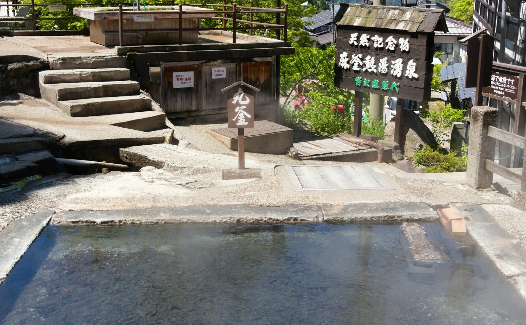 A steaming outdoor hot spring bath at a traditional Japanese onsen, empty, with stone steps and wooden signage